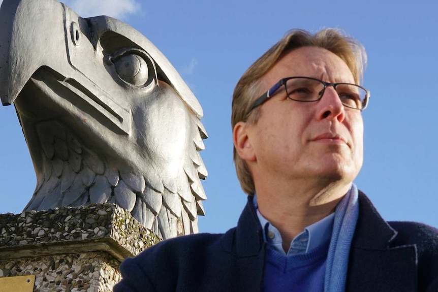 A man with fair hair glasses sands by a sculpture of an eagle.