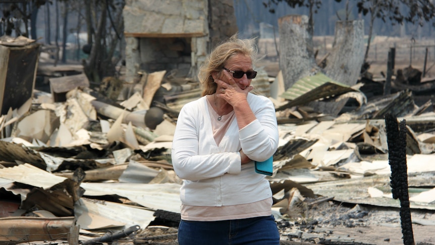 A woman holds rests her chin in her hands as she looks over damage cause by a bushfire.