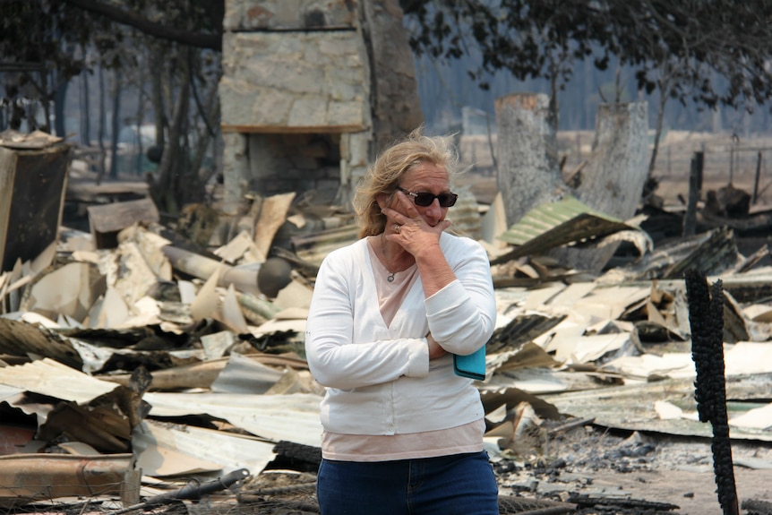 A woman holds rests her chin in her hands as she looks over damage cause by a bushfire.