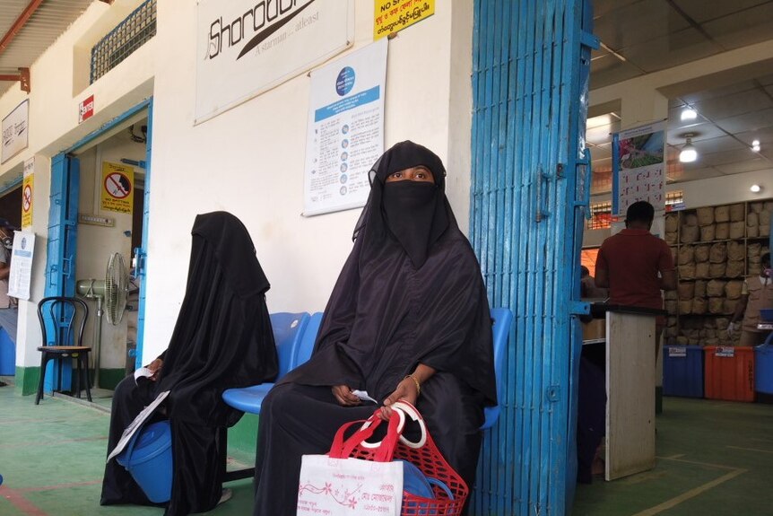 Two Muslim women in black abaya with face covering sit on a bench outside a shop.