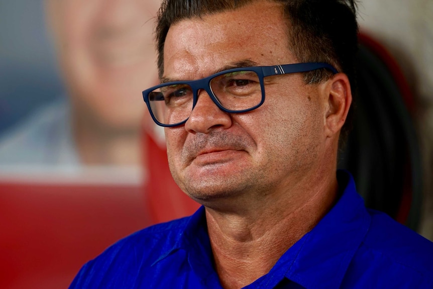 Ken Vowles, wearing a blue shirt, dark rimmed glasses and a concerned expression