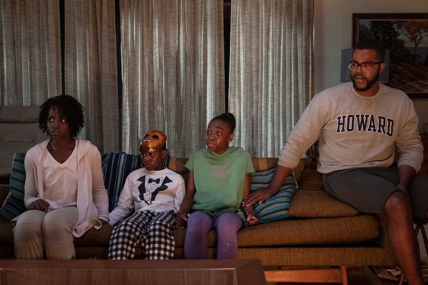 The actors are seated side by side on a living room couch, all look deeply troubled and the children have tear-stained faces.