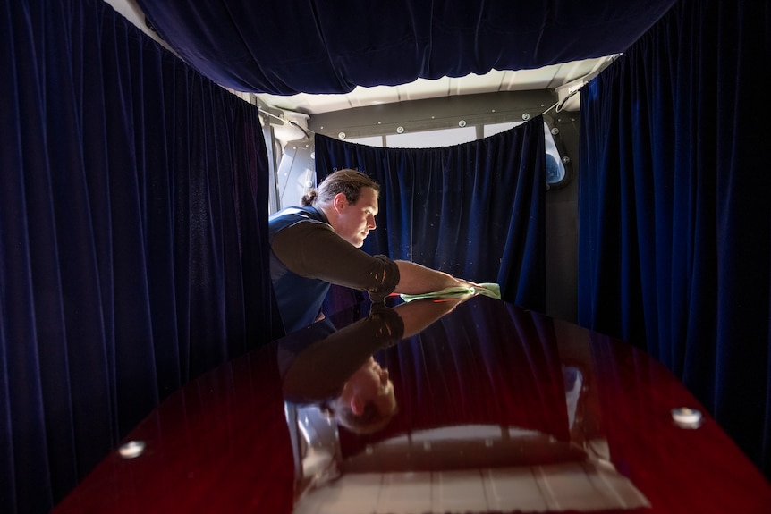 A adjusts a coffin that sits in a curtained hearse. His face reflects in the shiny wood of the coffin