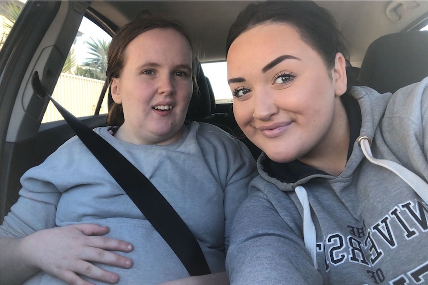 Two young women wearing grey tops smile at the camera as one of them takes a selfie in a car