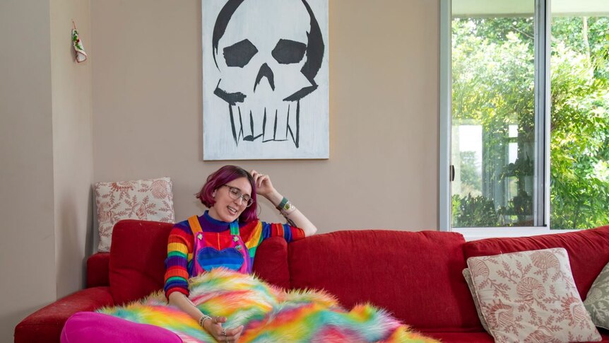 A woman with short pink hair sits on a red couch under a rainbow jumper under an artwork of a skull.