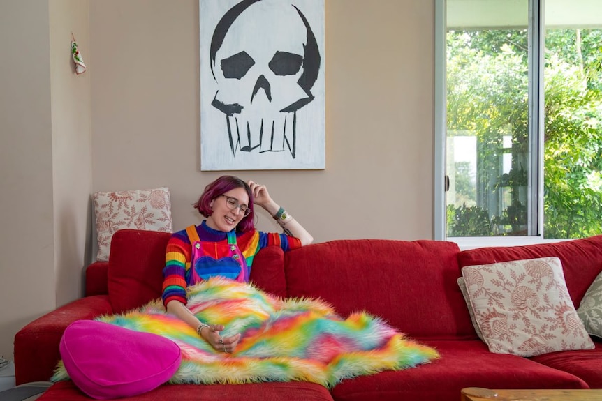 A woman with short pink hair sits on a red couch under a rainbow jumper under an artwork of a skull.