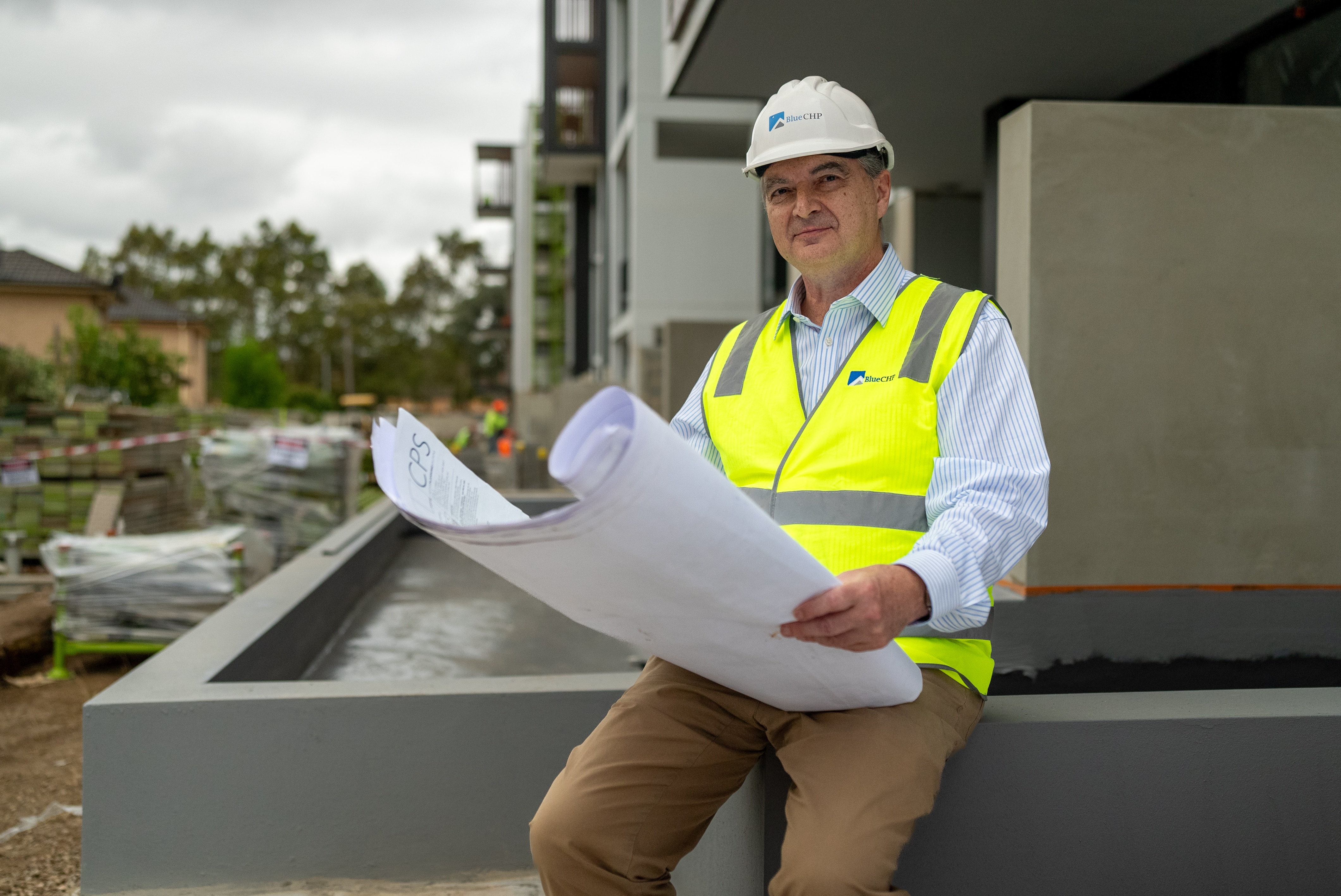 Hombre con camisa azul, chaleco de alta visibilidad y casco sosteniendo planos arquitectónicos.