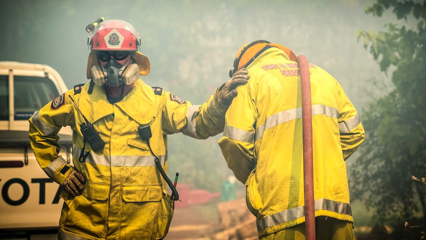 A mid shot of two firefighters in protective gear standing side by side with one of them resting a hand on the other's shoulder.