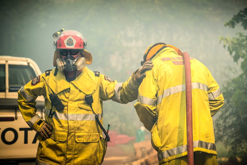 A mid shot of two firefighters in protective gear standing side by side with one of them resting a hand on the other's shoulder.