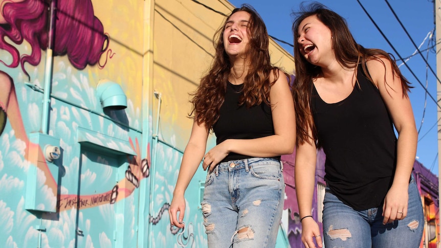Two young women with dark hair laugh while walking next to a brightly coloured wall.