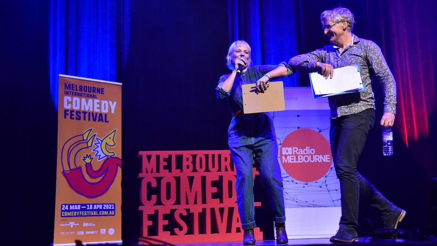 A woman and a man bump elbows on stage in front of banners for the Melbourne International Comedy Festival and ABC Radio Melb.