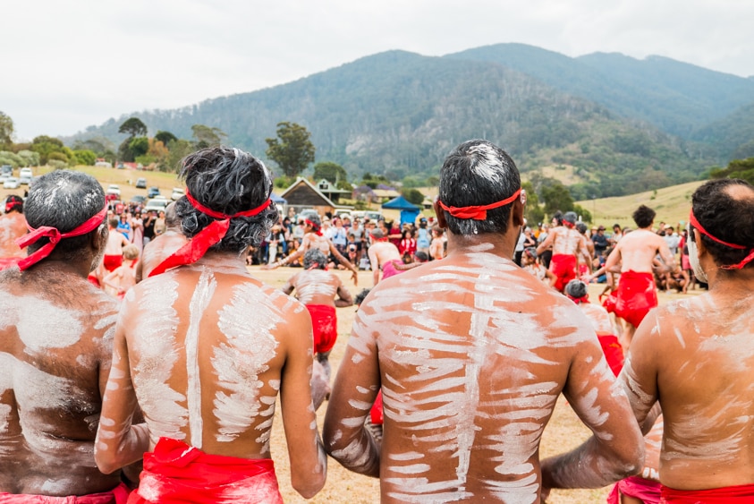 Four men in traditional Indigenous ceremonial dress and ochre watch as others dance, with Mount Gulaga in the background