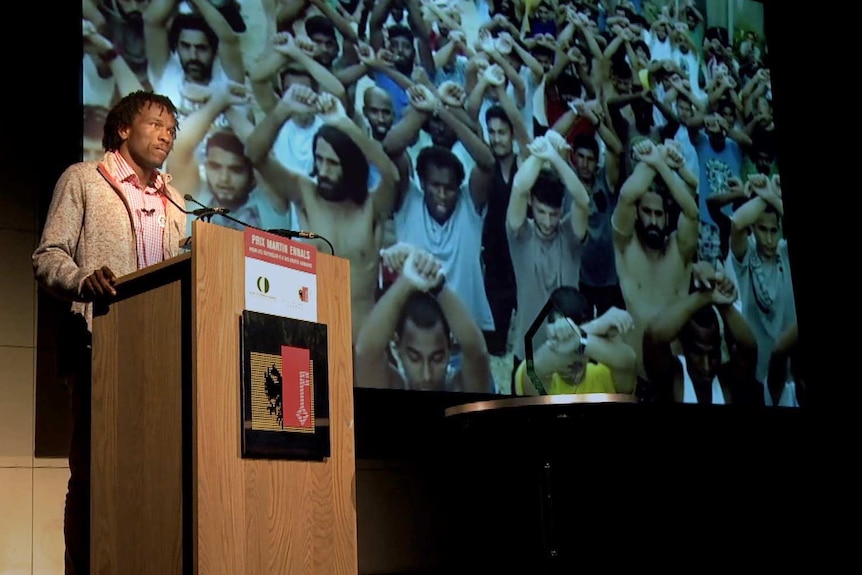 Man stands behind a lectern in front of a large screen showing people holding their hands up.