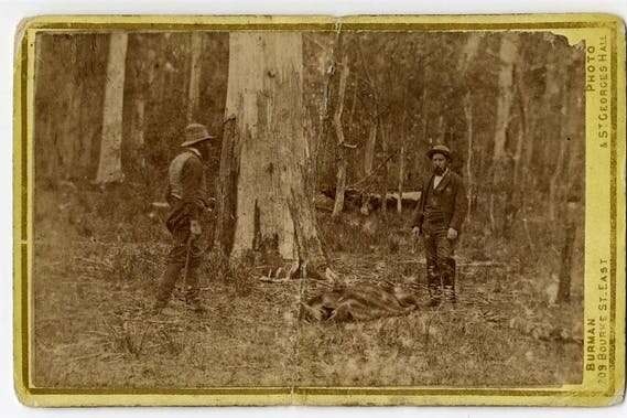 An old, sepia-toned photograph of two men standing near the body of another man.