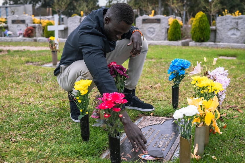 A man crouches and reaches out to touch a grave. The plaque of the grave is surrounded but colourful bunches of flowers.