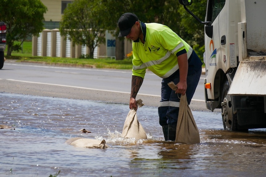 A man in high-vis lugs sandbags along a flooded road.