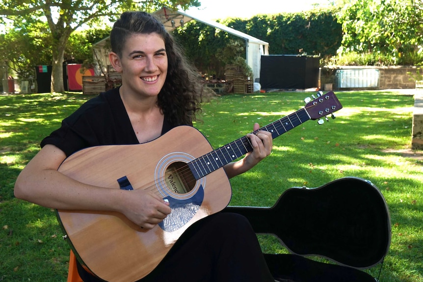 A woman wearing a black t-shirt and holding a guitar sits on a lawn.