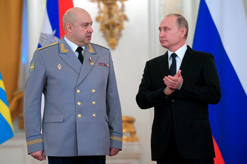 Sergei Surovikin wears a grey jacket as he stands next to a clapping Putin.