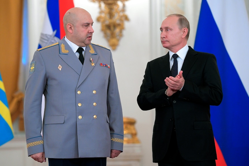 Sergei Surovikin wears a grey jacket as he stands next to a clapping Putin.