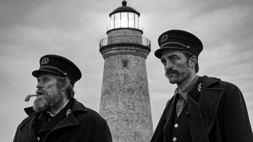 Black and white photo showing and older man and younger man in naval-type uniforms with a lighthouse in the background.