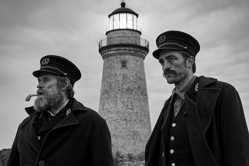 Black and white photo showing and older man and younger man in naval-type uniforms with a lighthouse in the background.