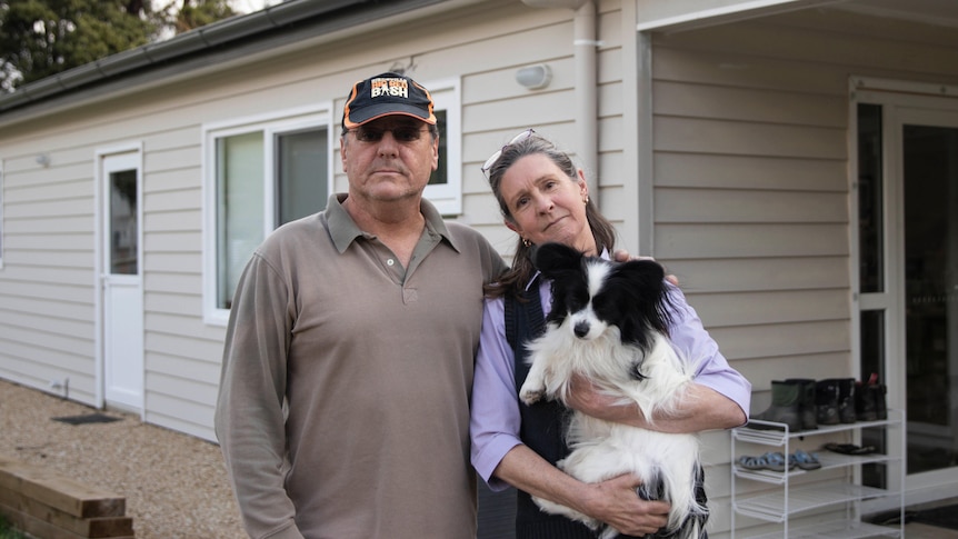 A man and a woman holding a dog stand together outside a house.