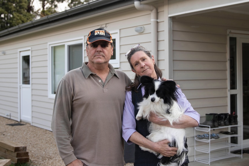 A man and a woman holding a dog stand together outside a house.