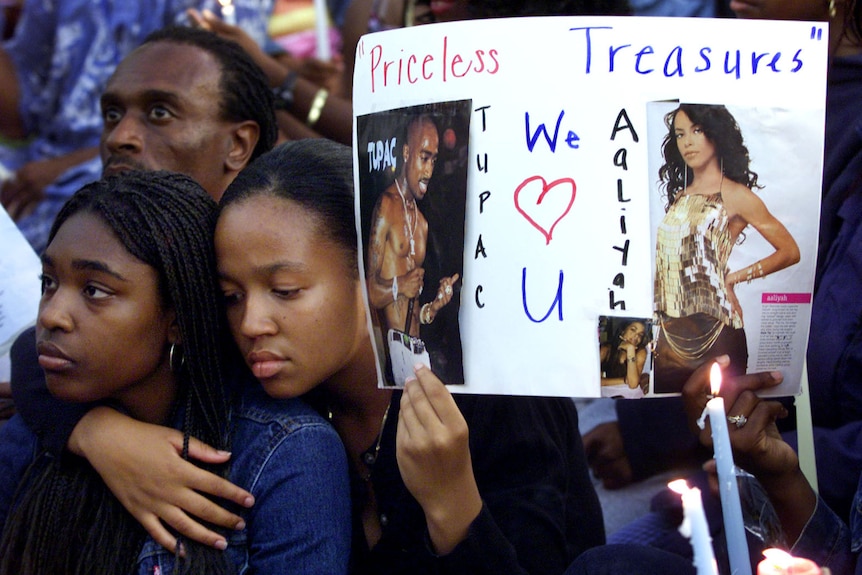 Two young black women attend a vigil, one of them holding a sign calling out Tupac and Aaliyah. "priceless treasures."