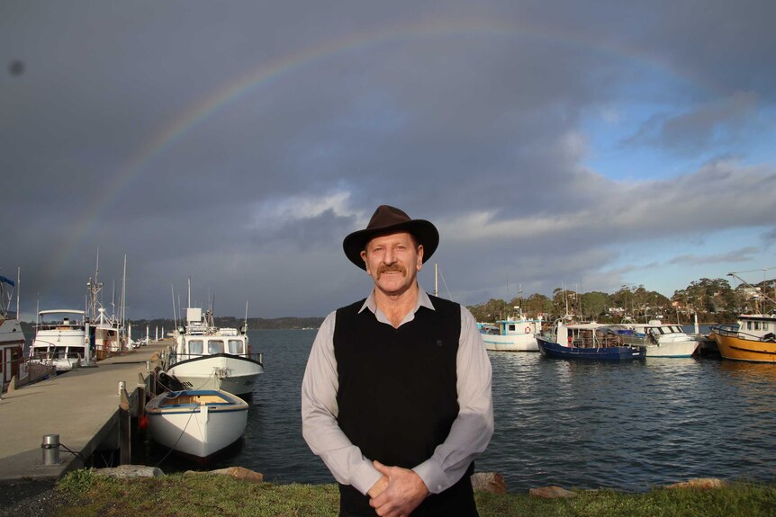 Man with a hat standing in front of dark grey clouds, rainbow and boats at the St Helens wharf