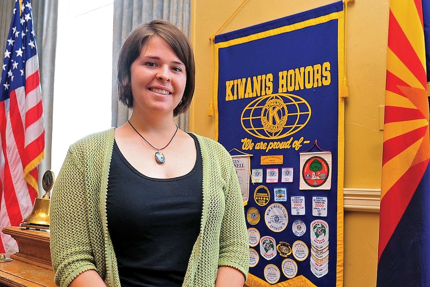 A woman with short brown hair stands in front of a US flag with her hands folded in front of her.