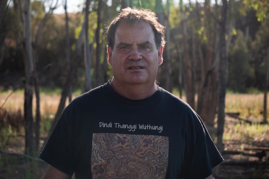 a man in a black shirt with traditional artwork and text that reads 'dindi thanggi wuthangi' is in the centre of the frame