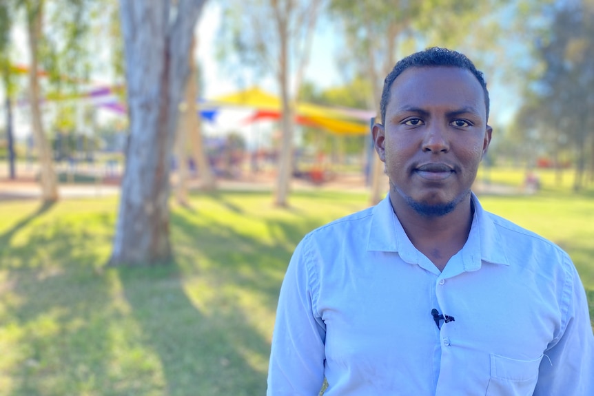 A Somalian man stanidng in a park wearing a white button up shirt. 