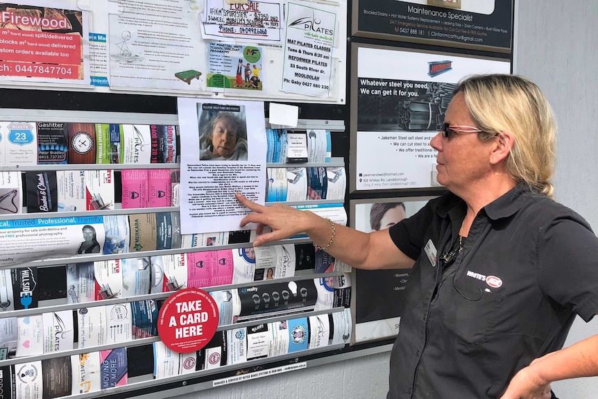 A woman standing next to a community notice board looking at a flyer on the elderly woman.