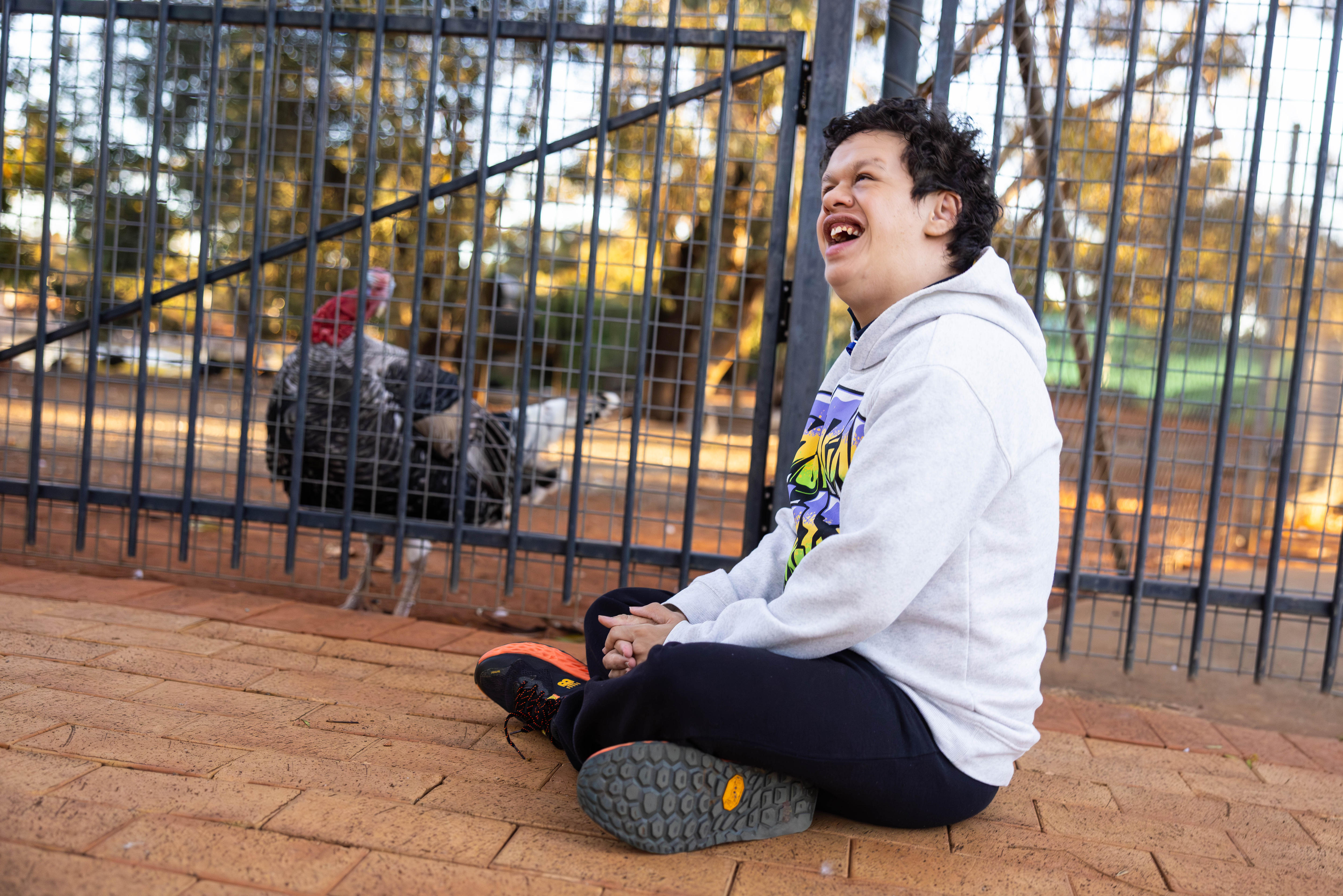 A teenage boy sitting down with a turkey behind fence.  