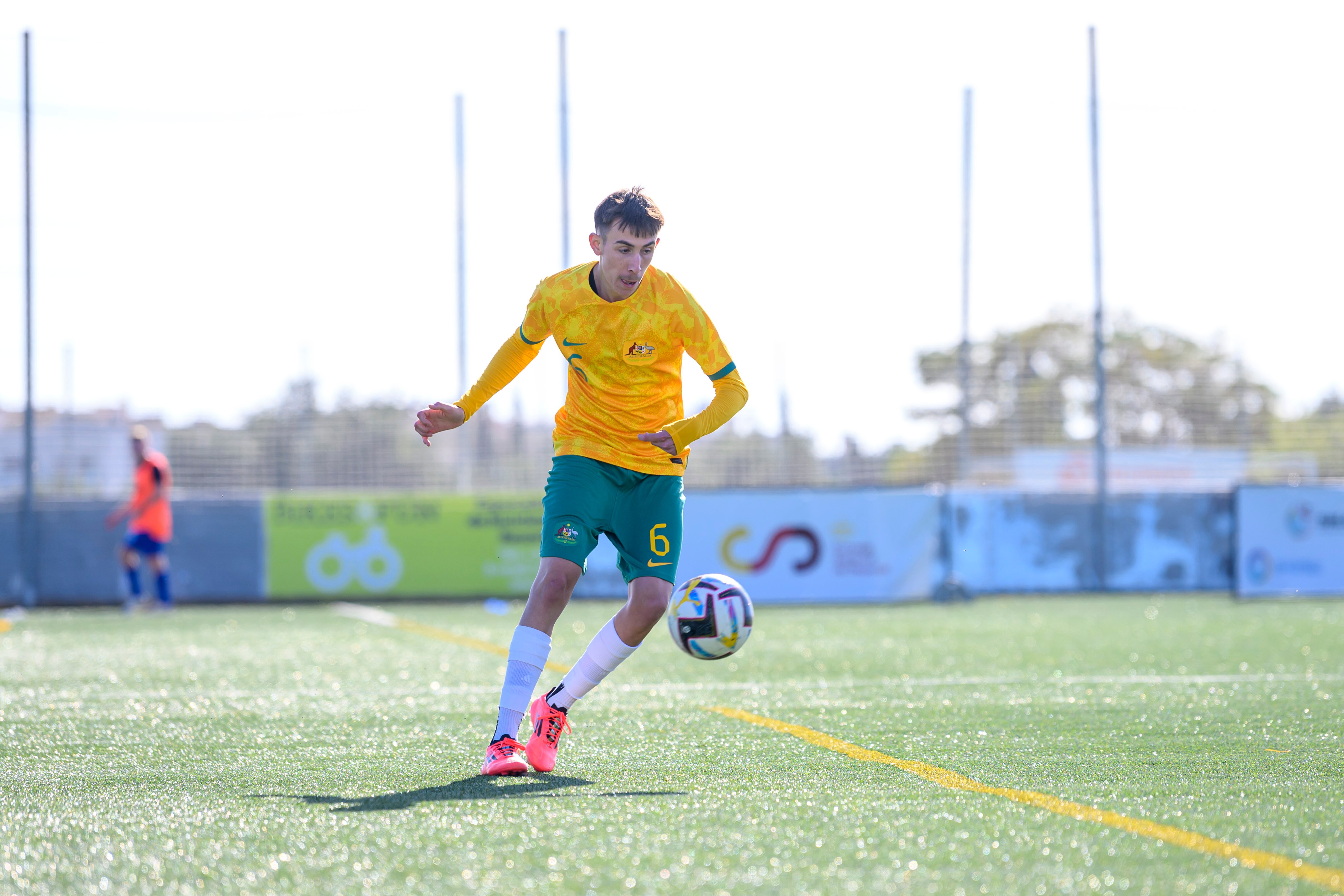 A man in a green and gold soccer uniform, white socks and pink soccer boots controls a ball during a match. He is on grass.