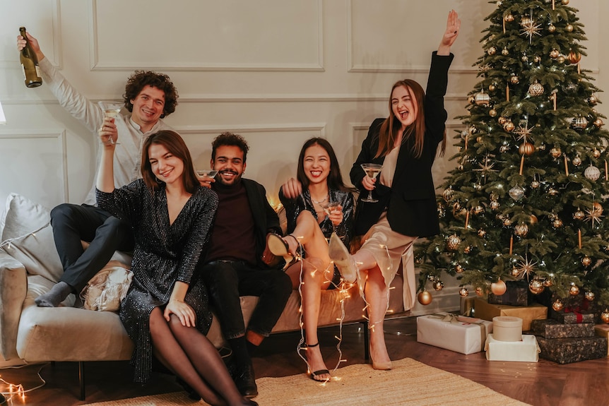 five friends sit on a couch next to a christmas tree with champagne in hand