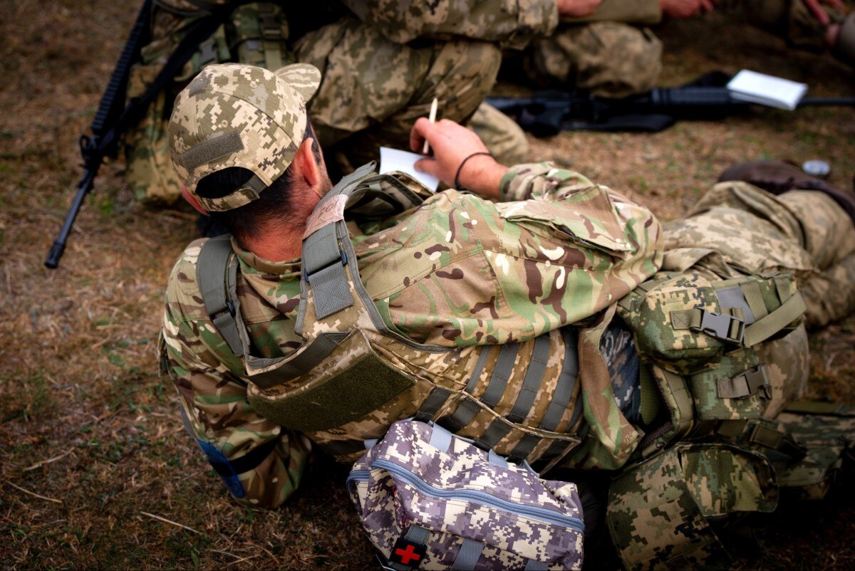 A soldier lying on the ground, writing something down, seen from behind.
