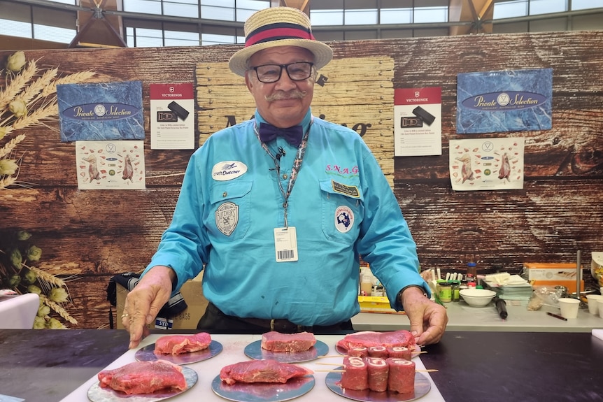 A man, who is wearing a blue short, top hat and bow tie, standing behind a table of beef steaks.