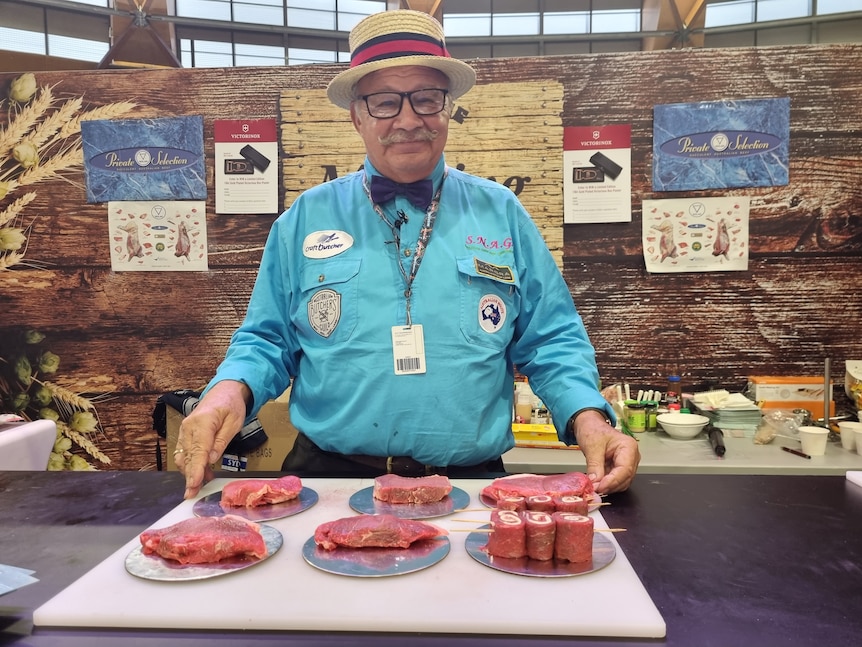 A man, who is wearing a blue short, top hat and bow tie, standing behind a table of beef steaks.