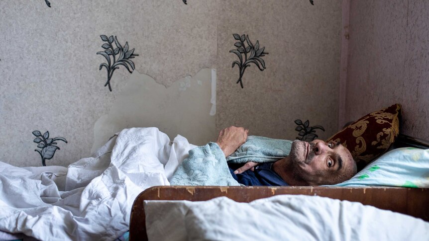A man lies on a bed covered in a blanket and towel with torn wallpaper behind him.