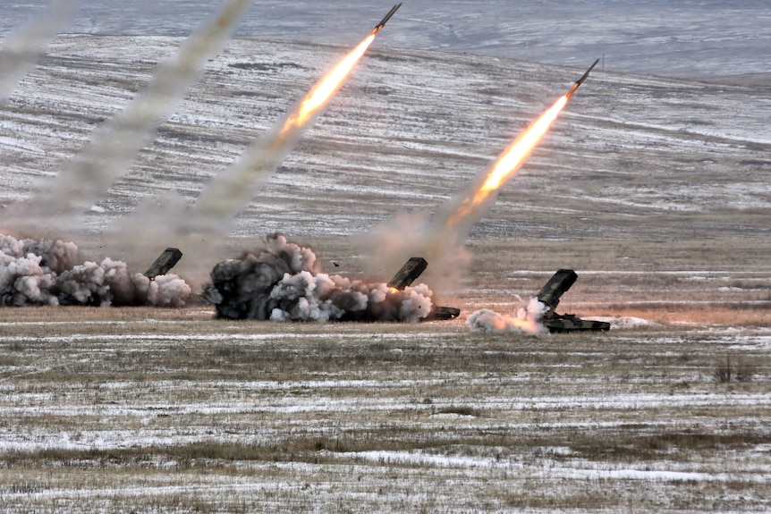 Rockets with long flaming tails fly through the air, with billowing clouds of black smoke wafting along the ground near tanks.