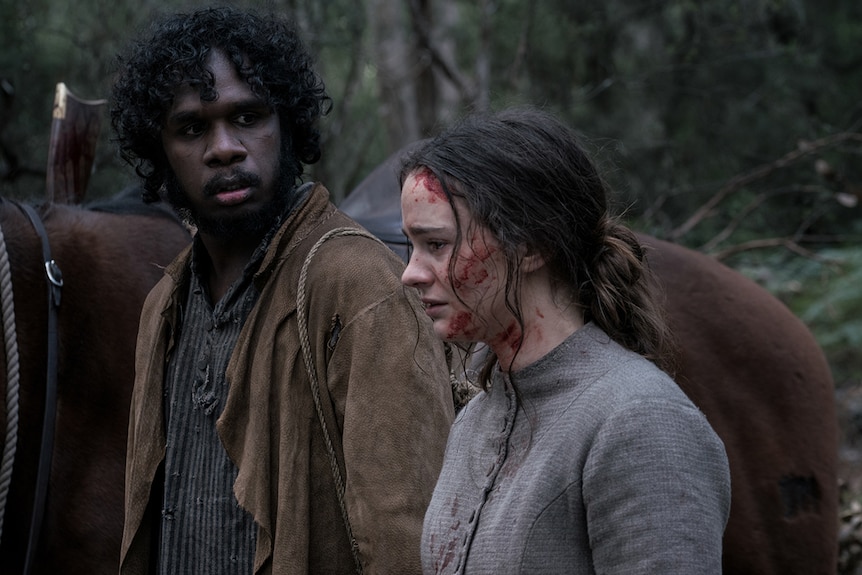 A man with worried expression looks towards a distraught woman covered in some blood, both in the bush.
