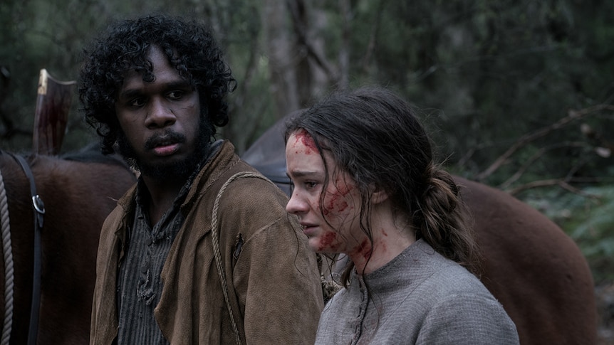 A man with worried expression looks towards a distraught woman covered in some blood, both in the bush.