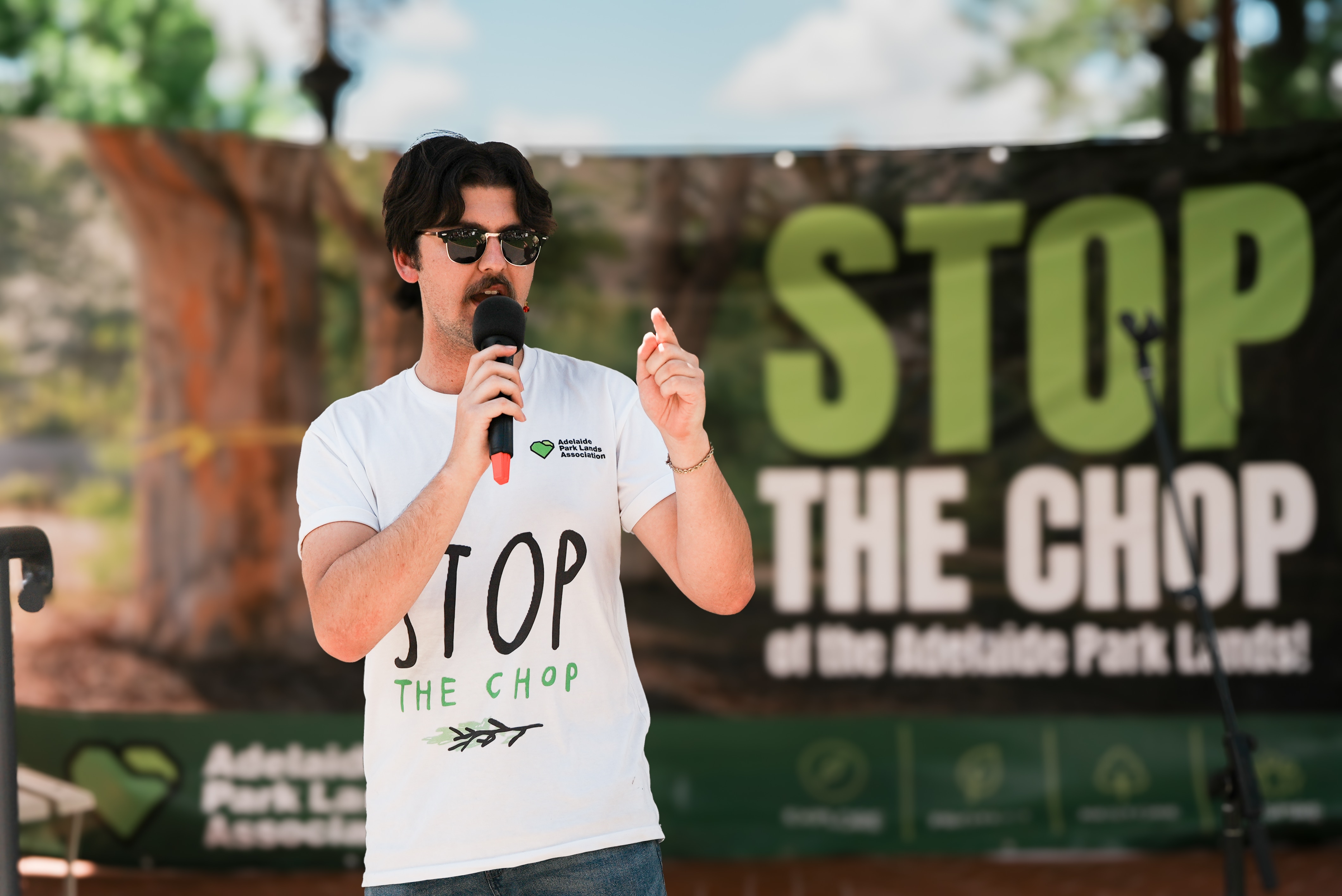 A man in front of a 'stop the chop' sign speaks on stage holding a microphone