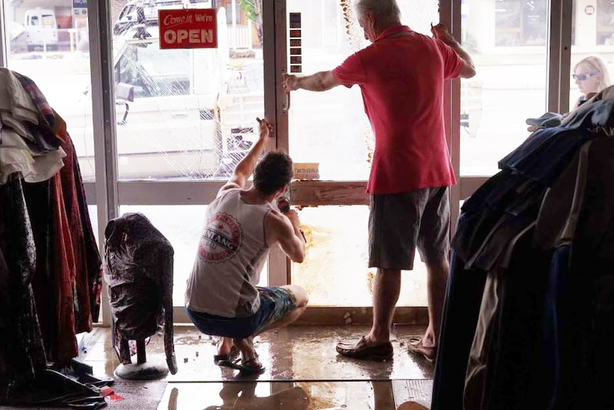 Two men tape up a broken plate glass window inside a menswear shop with racks of clothing behind them