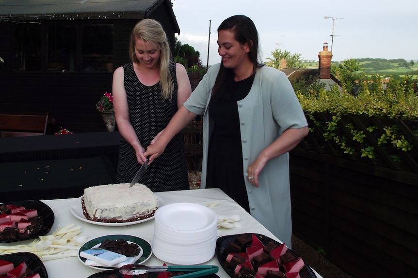 A woman with long black hair and another woman with long blonde hair cutting a cake on their wedding day. 