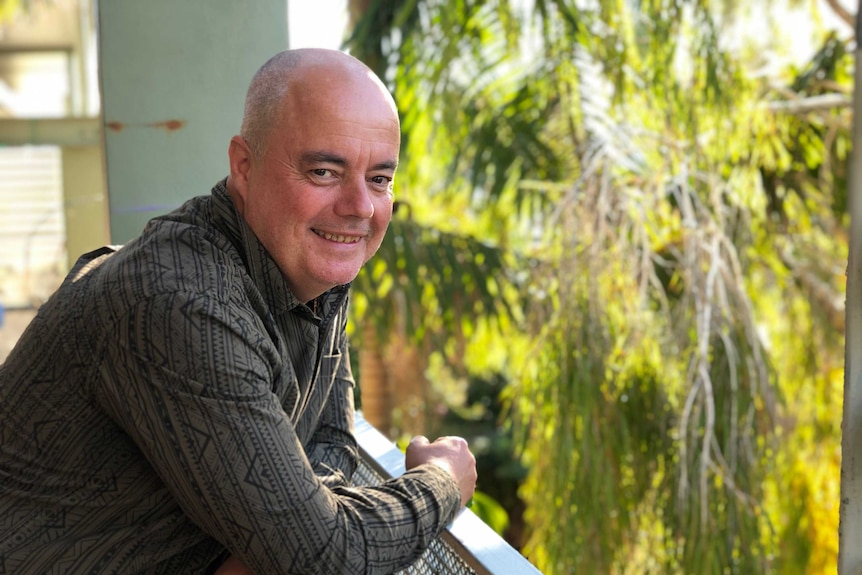 A man leaning over a rail smiling, with trees in the background.