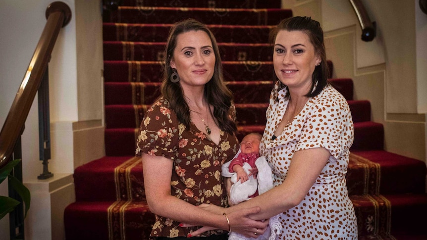 Two women smile at the camera and hold a baby