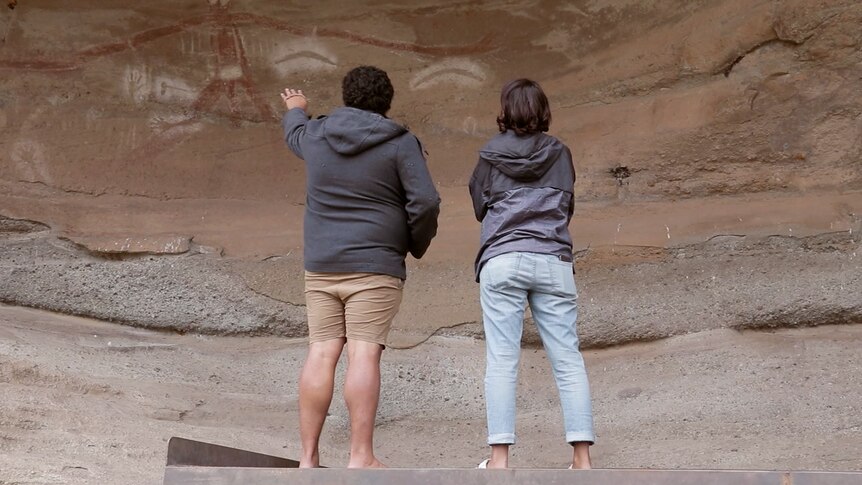 An Aboriginal man and woman stand on a platform overlooking an ancient cave site. The man points to markings inside the cave.