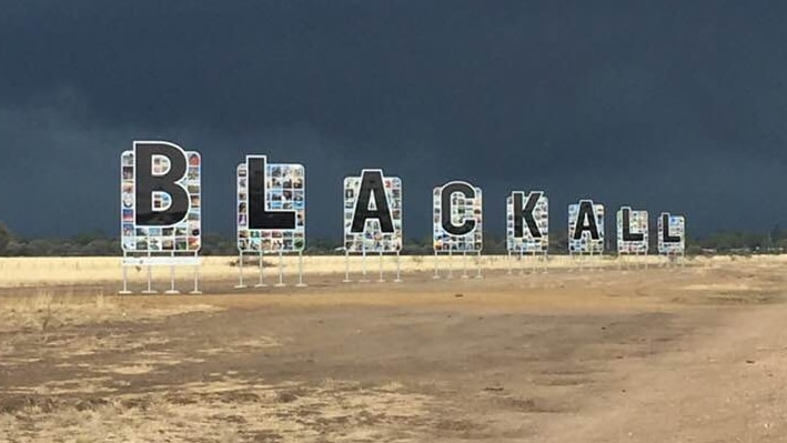 The letters B L A C K A L L on a sign in front of a dark sky full with storm clouds